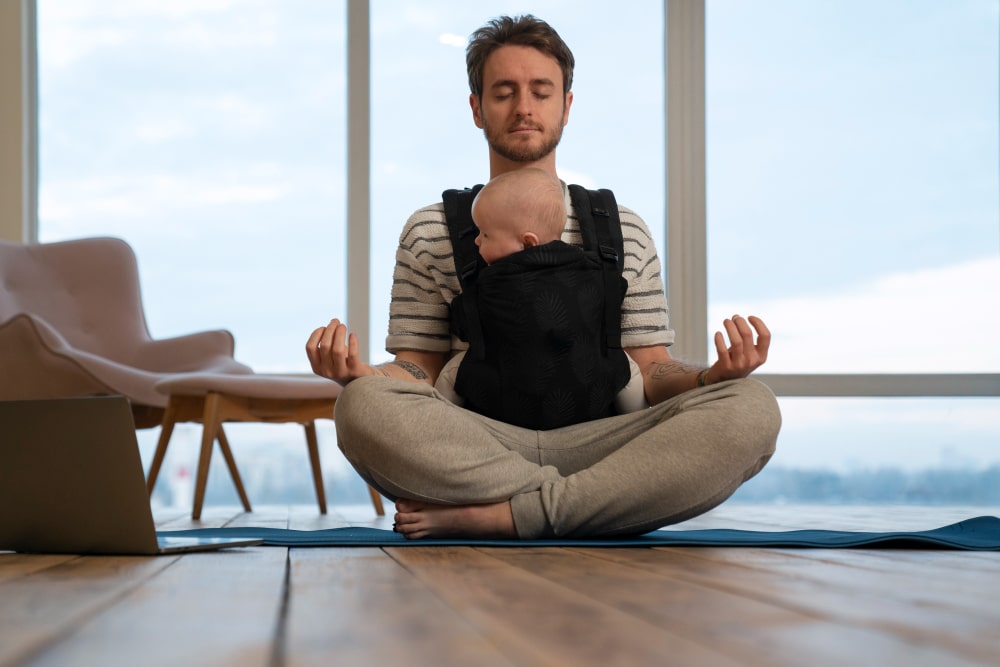 Jeune papa pratiquant une séance de sophrologie en ligne avec son bébé en portage pour apprendre à gérer la fatigue et le stress parental.